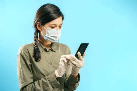 Young woman in protective medical mask and gloves using smartphone on a blue background. Online chat.の写真素材