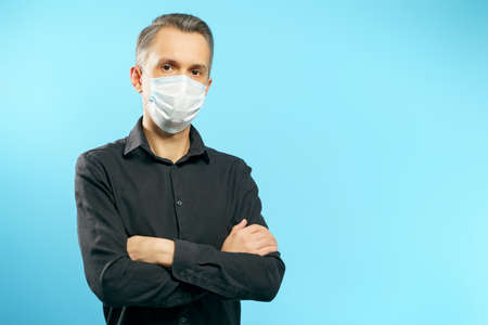 Portrait of a young man in a protective medical mask with arms crossed on a blue background.の写真素材