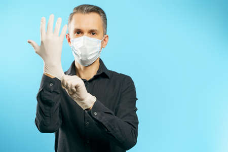 Portrait of a young man in a medical mask putting on protective gloves on a blue background.の写真素材