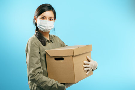 Young woman in medical protective mask and gloves holding a cardboard box on a blue background....