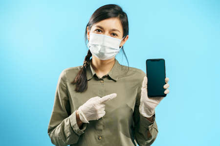 Portrait of a young woman in a protective medical mask and gloves pointing to the screen of a smartphone on a blue background. Copy space.の写真素材