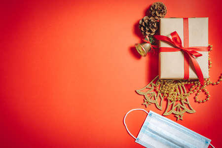 Composition of Christmas decorations, a gift box in a ribbon, fir cones and a protective medical mask on a red background.の写真素材