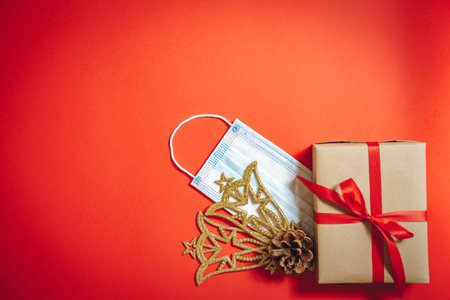 Composition of Christmas decorations, a gift box in a ribbon and a protective medical mask on a red background.の写真素材