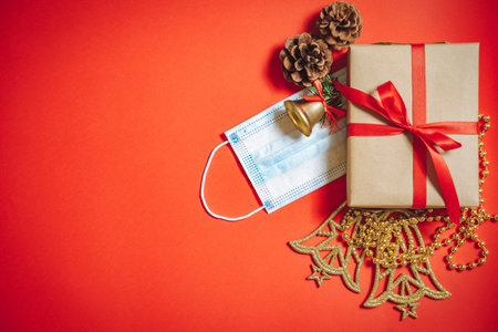 Composition of Christmas decorations, a gift box in a ribbon, fir cones and a protective medical mask on a red background. Copy space.の写真素材