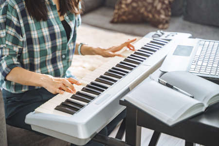 Close up of hands of unrecognizable young woman playing electric piano teaching remotely using laptop while working from home. Online education and leisure concept.の写真素材