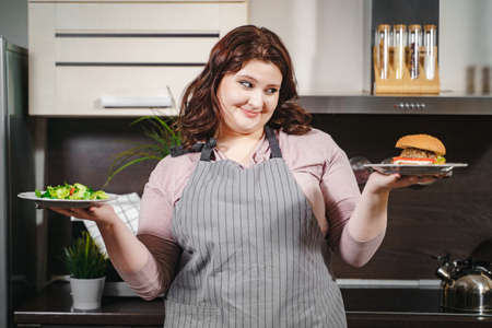 Portrait of a happy plus size woman holding a plate with burger and plate and salad in the kitchen. The concept of choosing between healthy nutrition and junk foodの写真素材