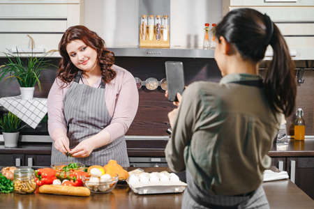 Two girlfriends recording a video or taking a photo on a smartphone for social networks. Plus size female blogger making blog content about diet and healthy food in the kitchen.の写真素材