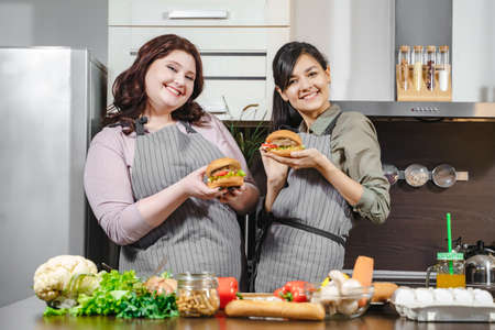 Two happy young women in aprons holding delicious burgers in their hands and looking at the camera in the kitchenの写真素材