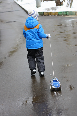 A small child (avid rear), which stands on the wet pavement and holds the rope toy car. The machine all in the snow. A child just walked through the snow.の写真素材
