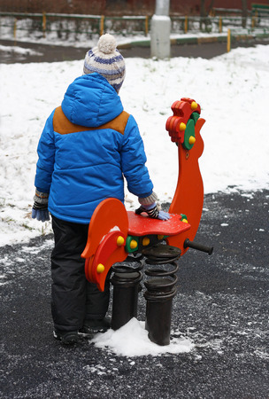 Walks in the fresh air. The image of the child in warm clothes (back view) standing on the Playground. Near children's rocking chair, made in the form of a bright red cock.の写真素材