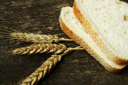 Ears of wheat on a brown wooden background. Near the ears are two cut pieces of white bread (rolls).