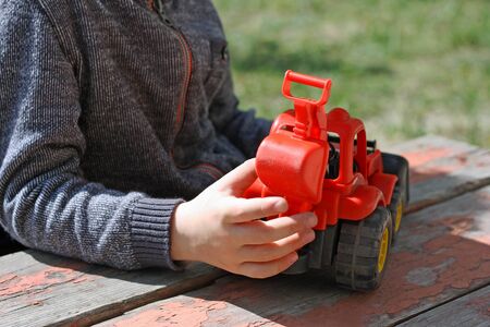 Image of a small child, who sits outdoors at the old table with wooden surface. A child plays with a red toy construction vehicles.の写真素材