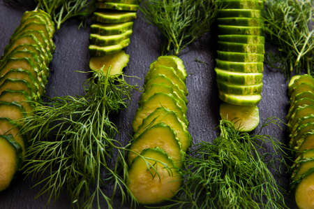 Natural fresh green cucumbers from a home garden on a black background, a dummy board made of stone. The cucumbers are cut into pieces and arranged in a dill pattern.の写真素材