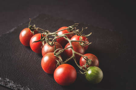 bunch of fresh cherry tomatoes on a branch are located on a black serving board on a dark backgroundの写真素材