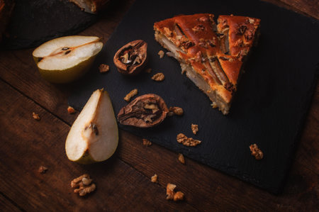 piece of pear pie on a black serving board on a wooden table, cut pears and walnuts lie chaoticallyの写真素材