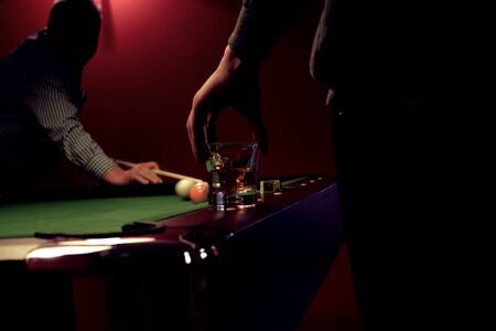 Pool billiard game. Close up of a whiskey glass in the hand of a billiard player. Billiard pool is in the backgroundの写真素材