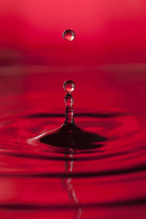 A red abstract background of a close up of transparent water drops falling in water, making beautifull ripples and flowing waves in the surface and showing the pure energy it creates. Small depth of field.の写真素材