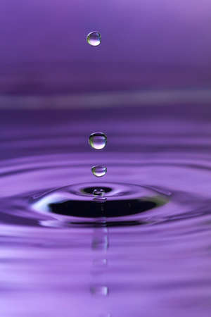 A purple abstract background of a close up of transparent water drops falling in water, making beautifull ripples and flowing waves in the surface and showing the pure energy it creates. Small depth of field.の写真素材