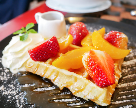 Closeup honey waffle with strawberry and peach and icing sprinkle on black plate (soft focus)の写真素材
