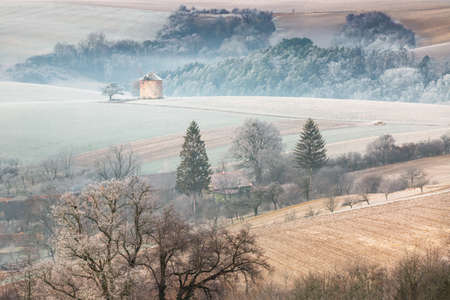 Winter rural landscape with frosted wavy fields, trees in hoarfrost and old windmill on hill. Moravia, Czech Republic.の写真素材