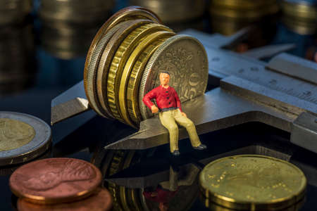Miniature people: Young man sitting on vernier caliper measuring coins stack. Retirement planning....