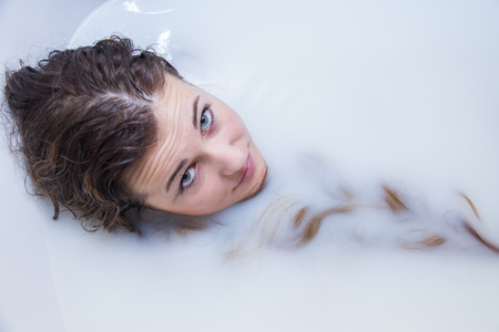 Portrait photo of a blonde girl with blue eyes bathing in a bathtub full of water and milk. She is...