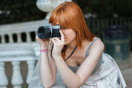 Young girl with orange hair taking pictures With an old camera retro style in a city park. She is...