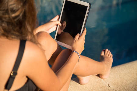 Side view of unrecognizable woman sitting and relaxing with digital tablet on poolside.の写真素材