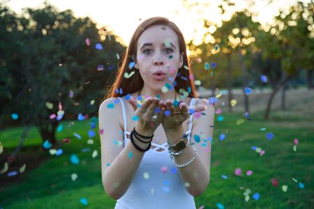 Pretty young woman standing in park on sunset and blowing confetti.