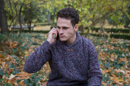 Young handsome man sitting on meadow with foliage and communicating with smartphone.の写真素材