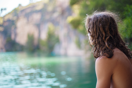 Young man looking at the greenish lake in the mountains at sunny day seen from his back.の写真素材