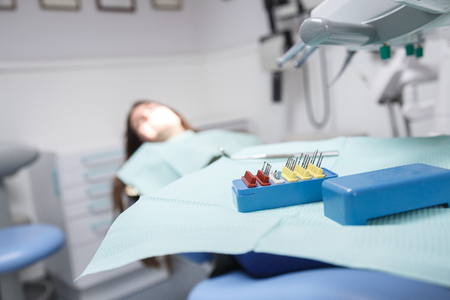Young female sitting in dental chair in clinic room in soft focus with equipment and instruments for cleaning the mouth and for removing cavities on focus.の写真素材