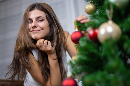 Beautiful young woman smiling and looking at camera while hanging small bauble on Christmas treeの写真素材