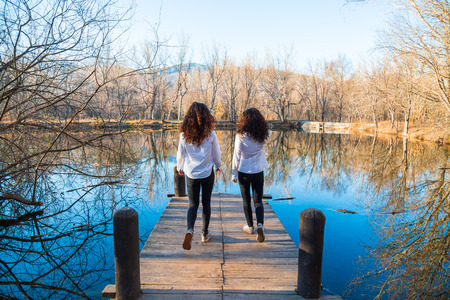 Back view of young twin sisters in the same clothes walking on pier on autumn day in the forestの写真素材