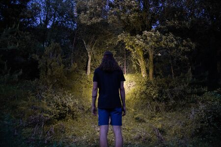 Back view of young man wearing shorts and t-shirt and illuminating forest with head lamp on summertimeの写真素材