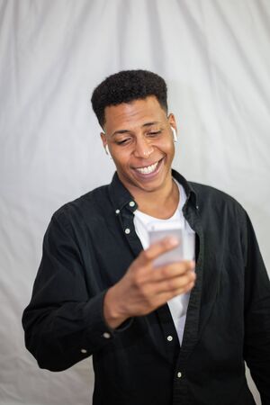 Happy african american young handsome man with wireless earphones in black shirt holding smartphone and laughingの写真素材