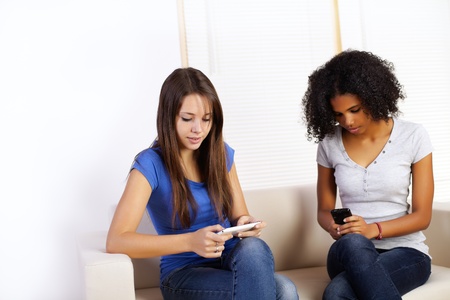 Portrait of two cute girls using mobile phones on a sofa.の写真素材