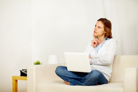 Portrait of a young adult woman thinking on sofa and holding a laptop while restingの写真素材