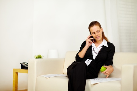 Portrait of a young caucasian business woman working with documents while is eating a green apple and speaking on the phoneの写真素材