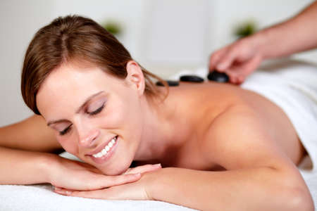 Close up portrait of a beautiful blonde woman smiling with her eyes closed while getting a hot stone massage from a professional masseuse at spaの写真素材