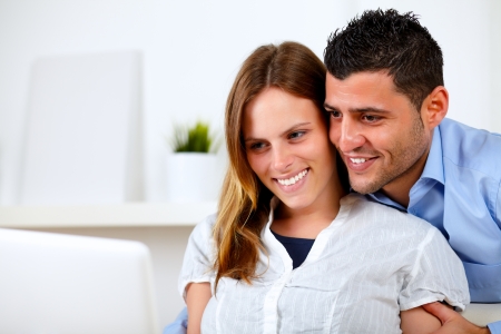 Portrait of a romantic young couple sitting on sofa at home looking to laptop screenの写真素材