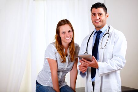 Portrait of a young pretty patient woman with her medical doctor looking at you at hospital indoorの写真素材