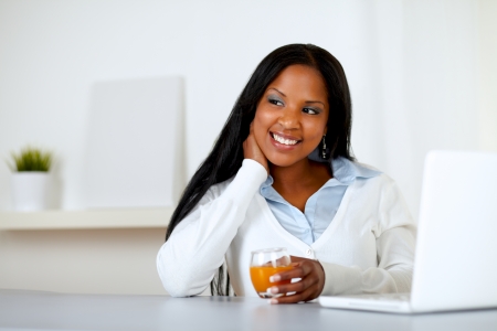 Portrait of an afro-american young woman holding an orange juice glass while thinking in front a laptopの写真素材