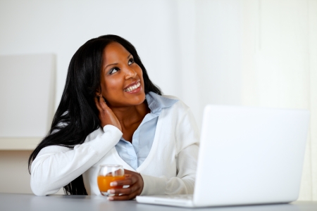 Portrait of an afro-american young woman looking up while is drinking an orange juice an having fun on laptop computer at homeの写真素材