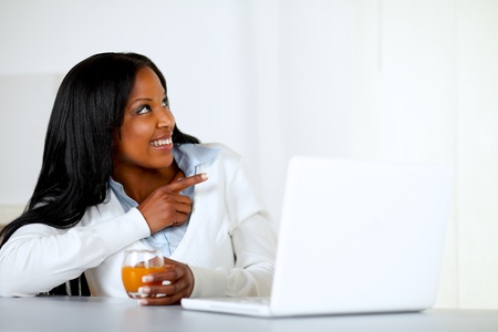 Portrait of an afro-american young woman drinking orange juice while is looking and pointing to her left in front a laptop at home indoorの写真素材