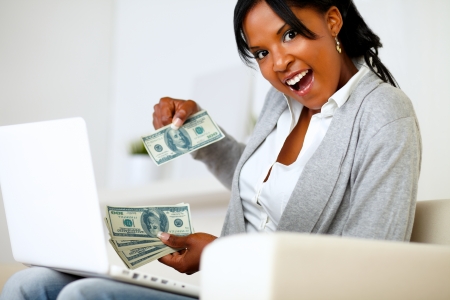 Portrait of a surprised woman with dollars is looking to you while is sitting on sofa at home in front a laptopの写真素材
