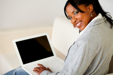Portrait of a relaxed afro-American female browse the Internet on laptop while is sitting on sofa at home indoor and is looking to youの写真素材