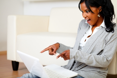 Portrait of an excited stylish woman pointing and looking to laptop screen while is sitting on the floor at homeの写真素材