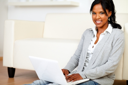 Portrait of a relaxed young woman smiling at you while is sitting on the floor using her laptop at home indoorの写真素材