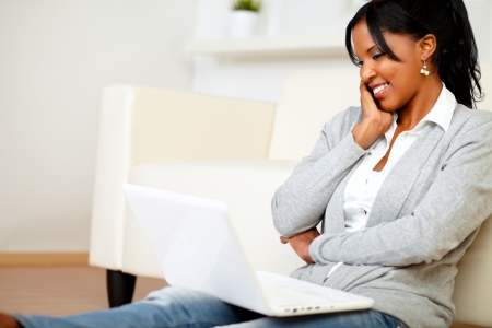 Portrait of a charming young woman smiling and looking to laptop while is sitting on the floor at home indoorの写真素材